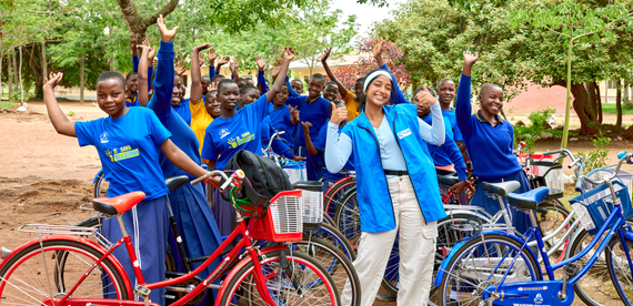 A young woman wears a Plan International vest and cheers alongside young female students with their bicycles outside their school in Tanzania.