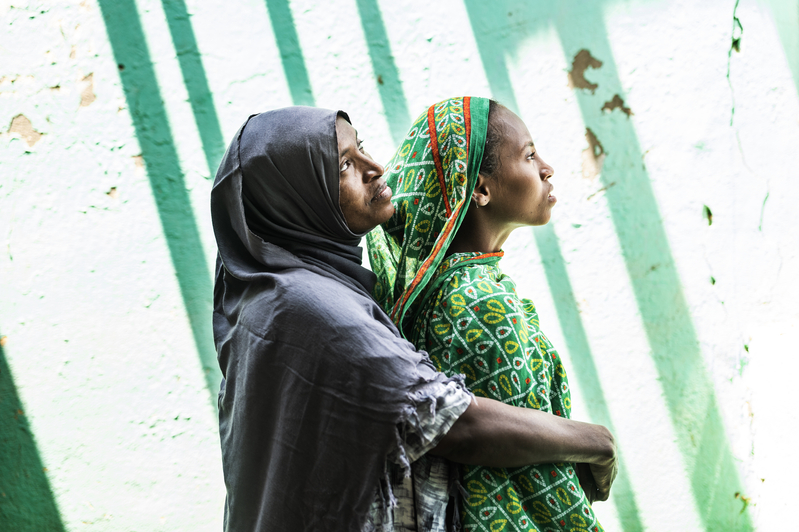 A woman in dark grey clothing wraps her arms around a younger woman in green from behind.