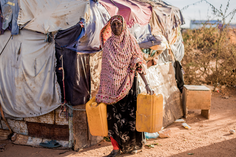 A woman in traditional attire carries two yellow jerry cans outside a makeshift tent in a dry, arid environment.