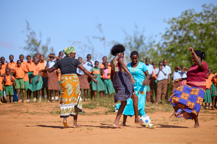 Four women are playing soccer in their bare feet on clay soil. There is a large group of girls and boys watching them play on the sidelines. 