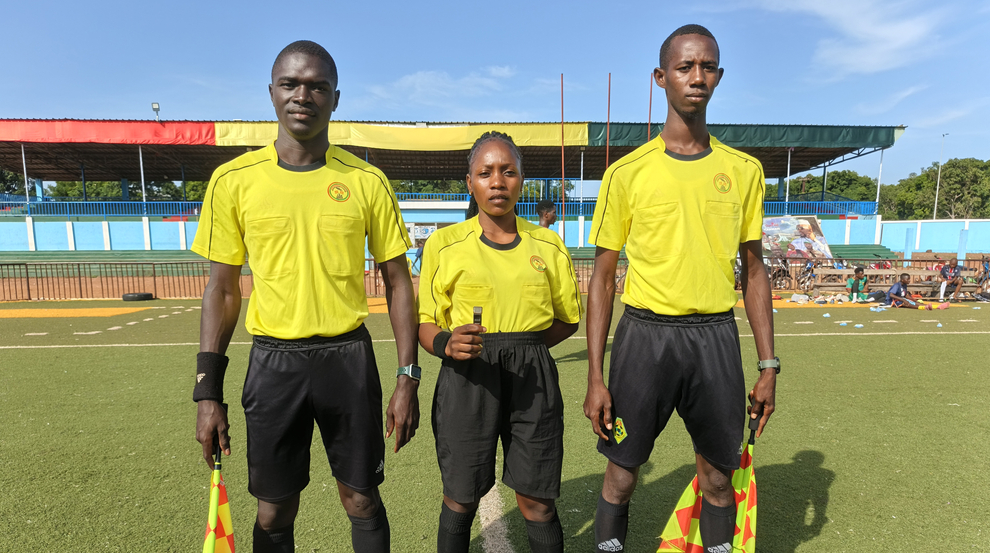 Three referees stand side by side on a football field. A woman wearing a yellow t-shirt and black shorts stands between two men wearing the same uniform. All three are facing the camera, with the field visible behind them.