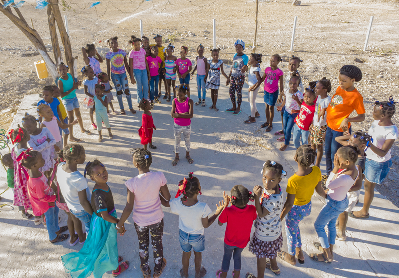 A group of children holding hands and standing in a circle outside. Two girls stand in the centre of the circle.