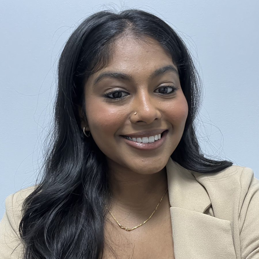 A young woman with long black hair in a beige blazer smiles at the camera in front of a light blue background.