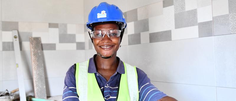 black girl wearing safty hat, glasses and vest smiling brightly inside a tiled room