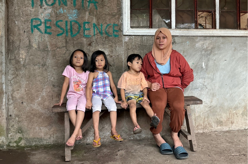 A woman and three young children sit together on a wooden bench outside a concrete building.