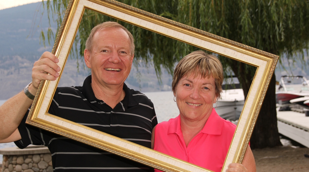 A couple holding a gold-coloured frame around themselves and taking a photo while standing outside near a lake.