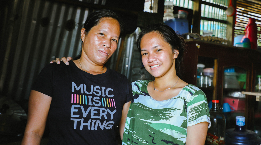 A woman in a black t-shirt and young girl in a green t-shirt stand facing the camera smiling. The young girl has her arm around the woman’s shoulder. 