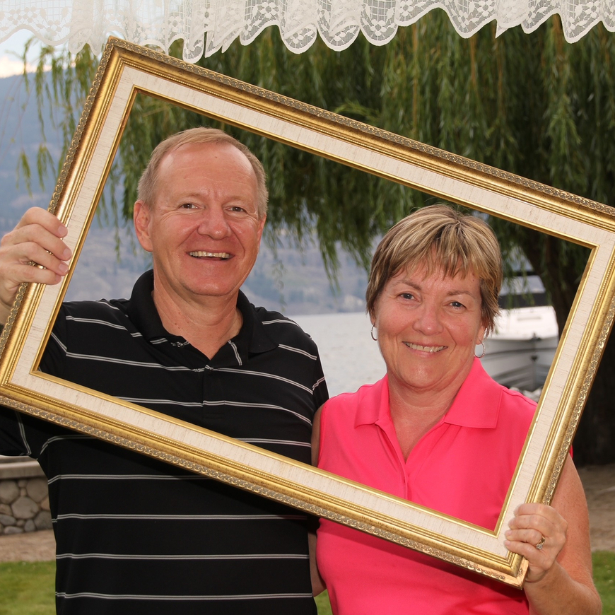 A couple holding a gold-coloured frame around themselves and taking a photo while standing outside near a lake.