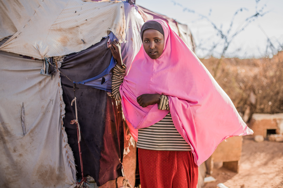A young girl in a pink shawl stands beside a makeshift tent, with a barren landscape in the background.