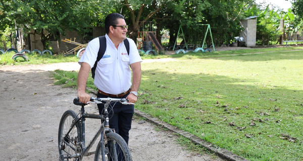 A man with dark hair and glasses standing on a dirt path, holding a bicycle near a grassy area with trees and a swing set in the background. 
