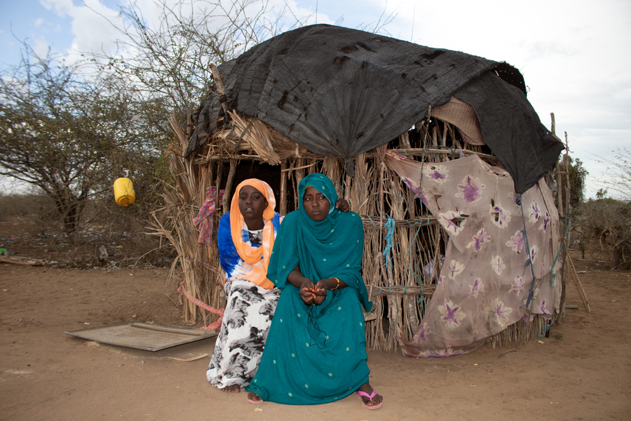 Two women sit outside a small, rustic hut made of sticks and fabric, surrounded by sparse vegetation in a dry landscape.