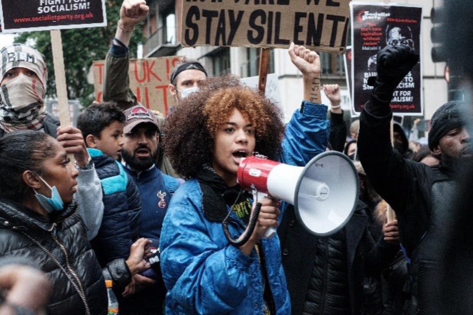 A woman speaks into a megaphone and is surrounded by people holding signs at a Black Lives Matter protest in 2020