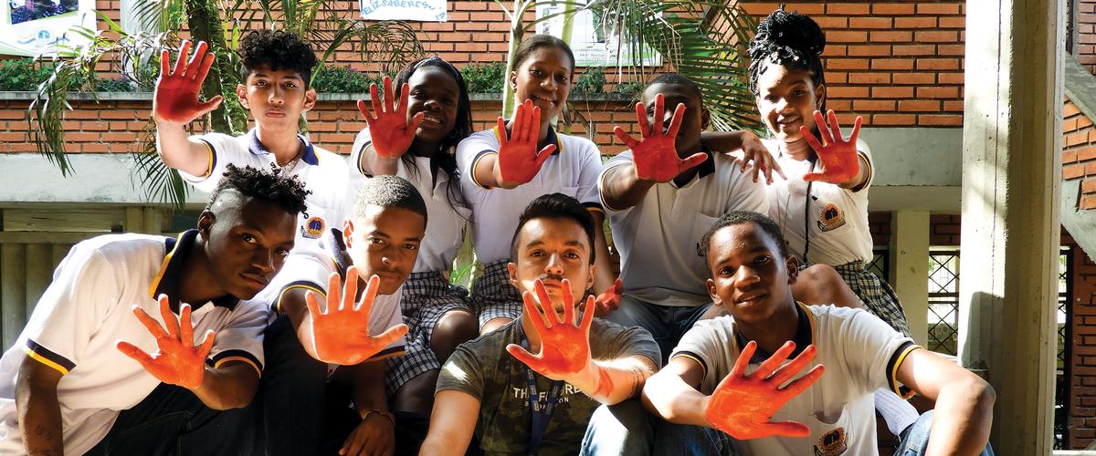 Nine young people sitting in a group holding up one hand each. Their hands are painted orange.