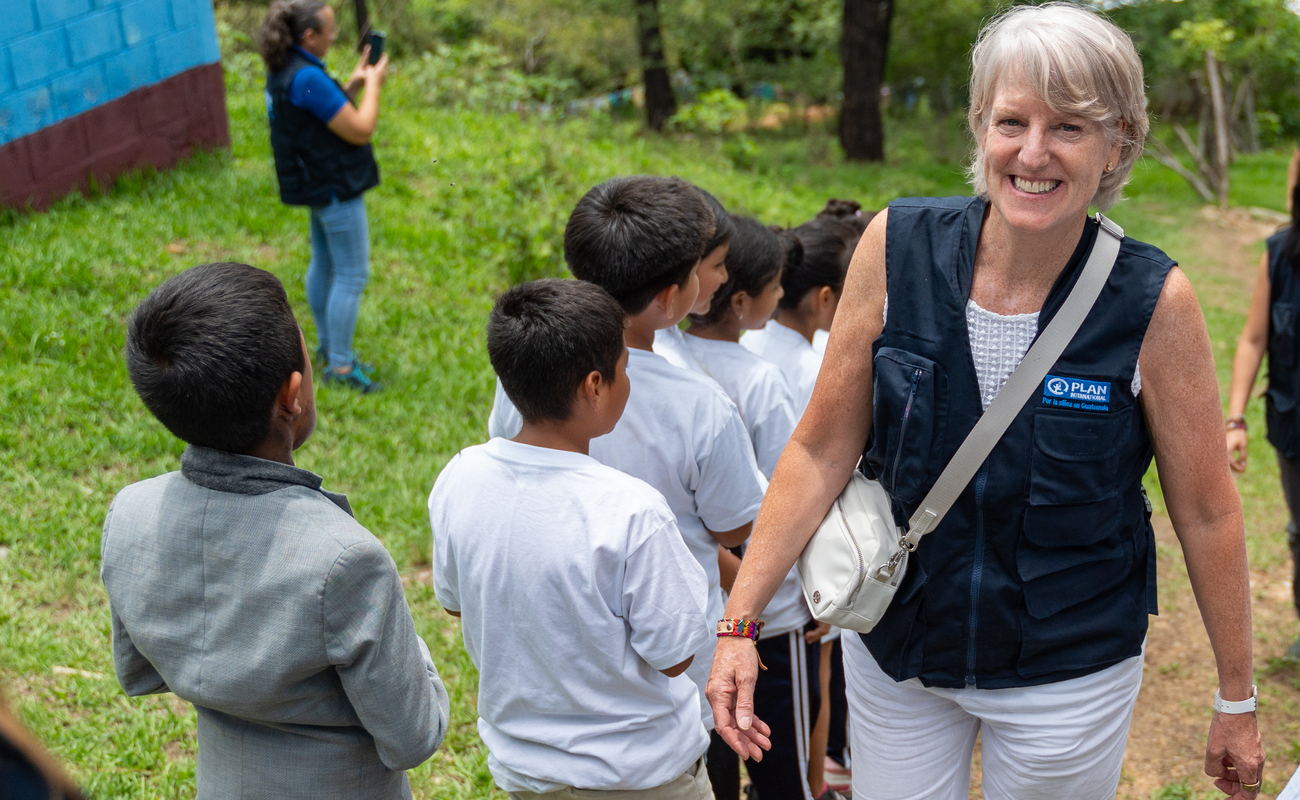 A woman with short blonde hair wearing a dark blue vest smiles with a group of children in white tshirts standing next to her in a line, with their backs to her.