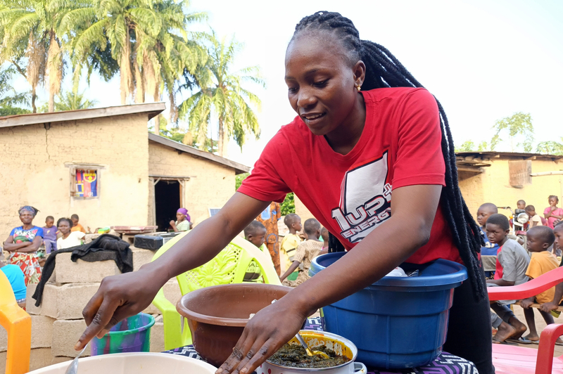 A woman in a red t-shirt stands in front of a large bowl of rice and other food outside while children and other community members sit in the background at tables.