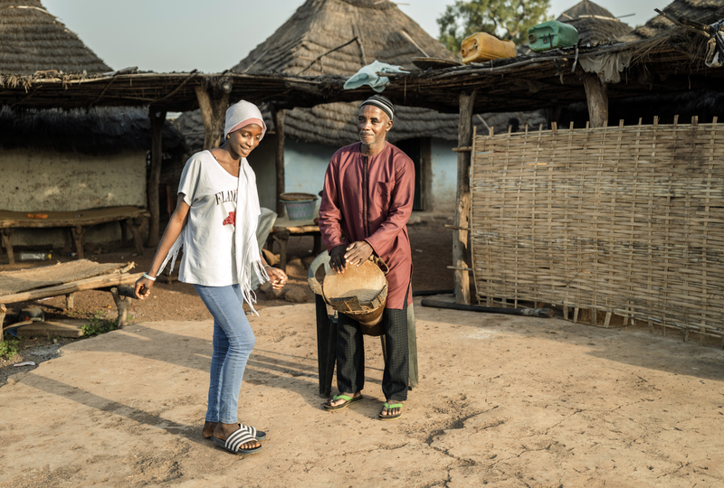 A father in Senegal wearing a red top and black pants plays a traditional drum while his teenage daughter wearing a white t-shirt and jeans dances beside him.