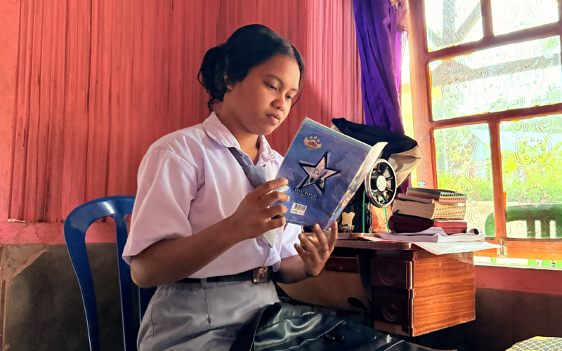 A student in a school uniform sitting by a window reading a book with textbooks nearby.