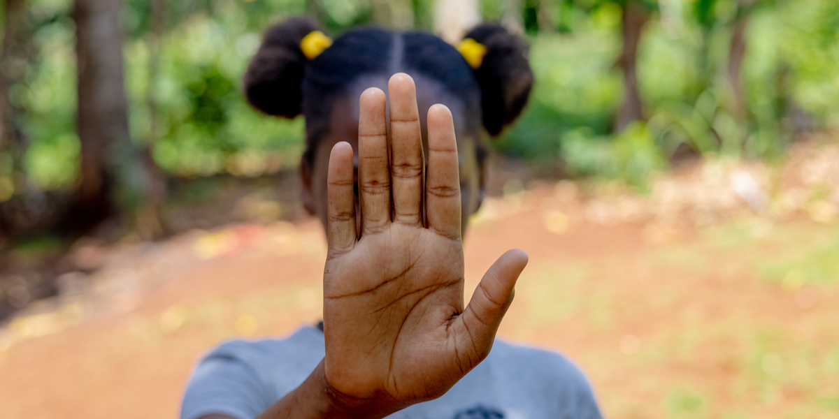 A girl with two buns in her hair holds up her hand, signaling a stop, against a blurred natural background.