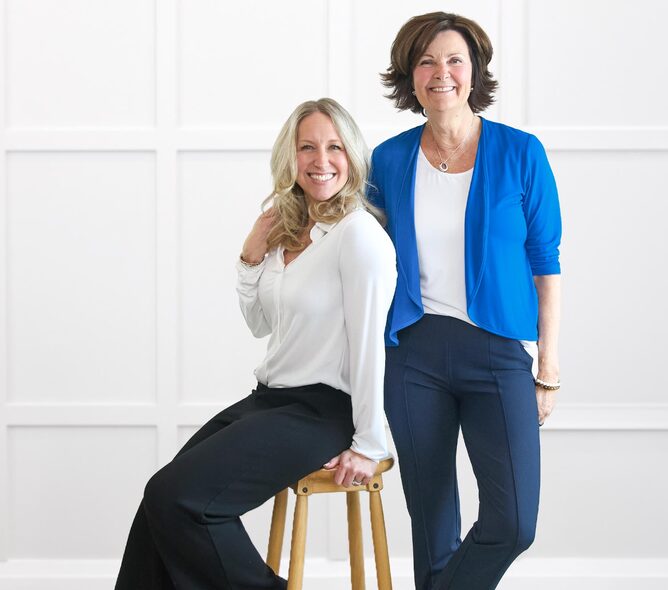 Two women smiling and posing together, one seated on a stool and one standing beside her