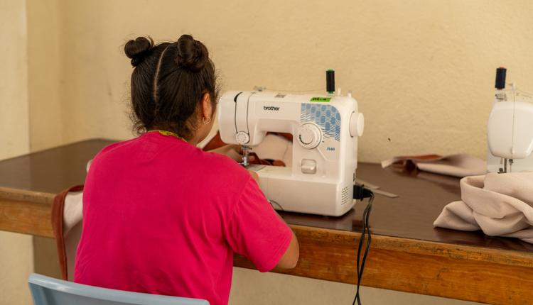 A young woman wearing a pink t-shirt works on a sewing machine.