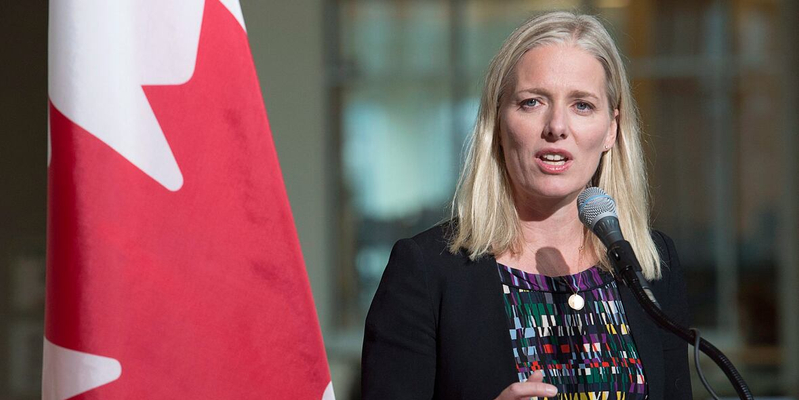 A woman speaks at a podium with a microphone beside a Canadian flag.  She is wearing a dark blazer over a patterned top and is addressing an audience in an indoor setting