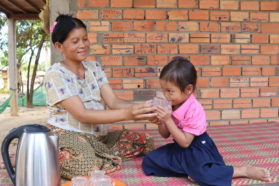 A girl holds a glass of water with the help of a woman who is smiling; they both sit on the ground on a mat while there is a kettle and other cups.
